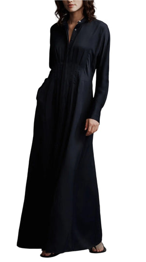 A woman stands against a white background wearing the TWP Main Street After Midnight dress—a long sleeve, floor-length black midi house dress featuring a buttoned neckline and side pockets.