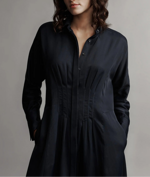 A woman stands against a neutral background wearing TWP's Main Street After Midnight—a long sleeve, button-up black midi house dress with pleated details and pockets. Her face is partially out of frame.
