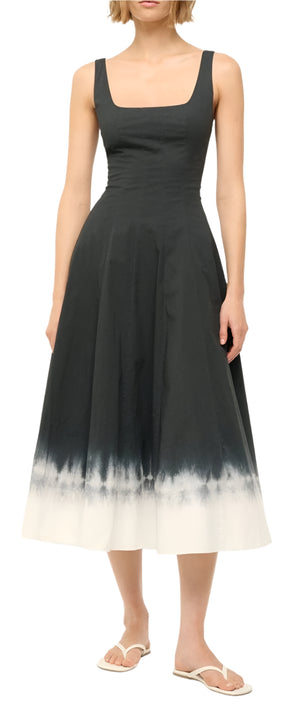 A person stands against a plain background in Staud's Wells Dress, a sleeveless black cotton poplin piece with a white gradient hem, complemented by chic white sandals for an elegant touch.