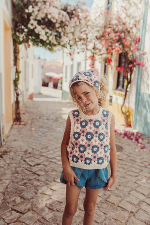 A young girl in the Louise Misha Girls' Rynia Vest stands on a cobblestone street with flowers and colorful buildings, wearing denim shorts and a matching headscarf over her curly blond hair.