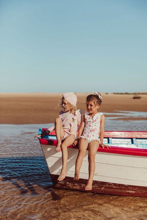 Two young girls wearing the Louise Misha Yanika Bathing Set by Louise Misha sit on a beached boat at the shore, with sand, shallow water, and a clear sky in the background.