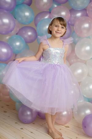 A young girl beams brightly as she stands barefoot before a backdrop of pastel balloons, wearing a glamorous Sequins Princess Dress by Great Pretenders in lilac.
