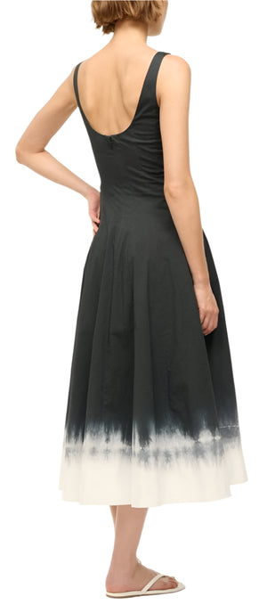 A woman is shown from behind, facing right, wearing Staud's Wells Dress, a sleeveless black and white tie-dye cotton poplin piece, against a plain background.