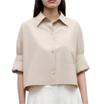 A woman wears TWP's Next Ex Vintage Washed Poplin, a beige cropped boxy button-up shirt with short sleeves and chest pocket, paired with white pants.
