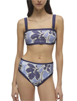 A woman in the Luthea Tube Top and Dallia Bottom by Simkhai, featuring a vibrant floral print, dark straps, and high-waisted bottoms with elegant satin fabric.