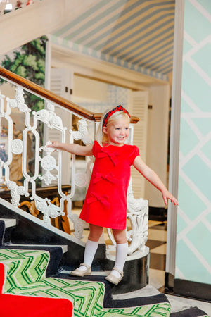 A young girl in a Girls' Velveteen Betts Bow Dress by The Beaufort Bonnet Company stands smiling on a staircase with patterned carpet and an ornate white railing.