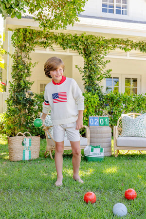 A barefoot boy in Boys' Prepletic™ Charlie's Chinos by The Beaufort Bonnet Company holds a green bocce ball on a grassy lawn, with bocce balls and a seating area in the background.