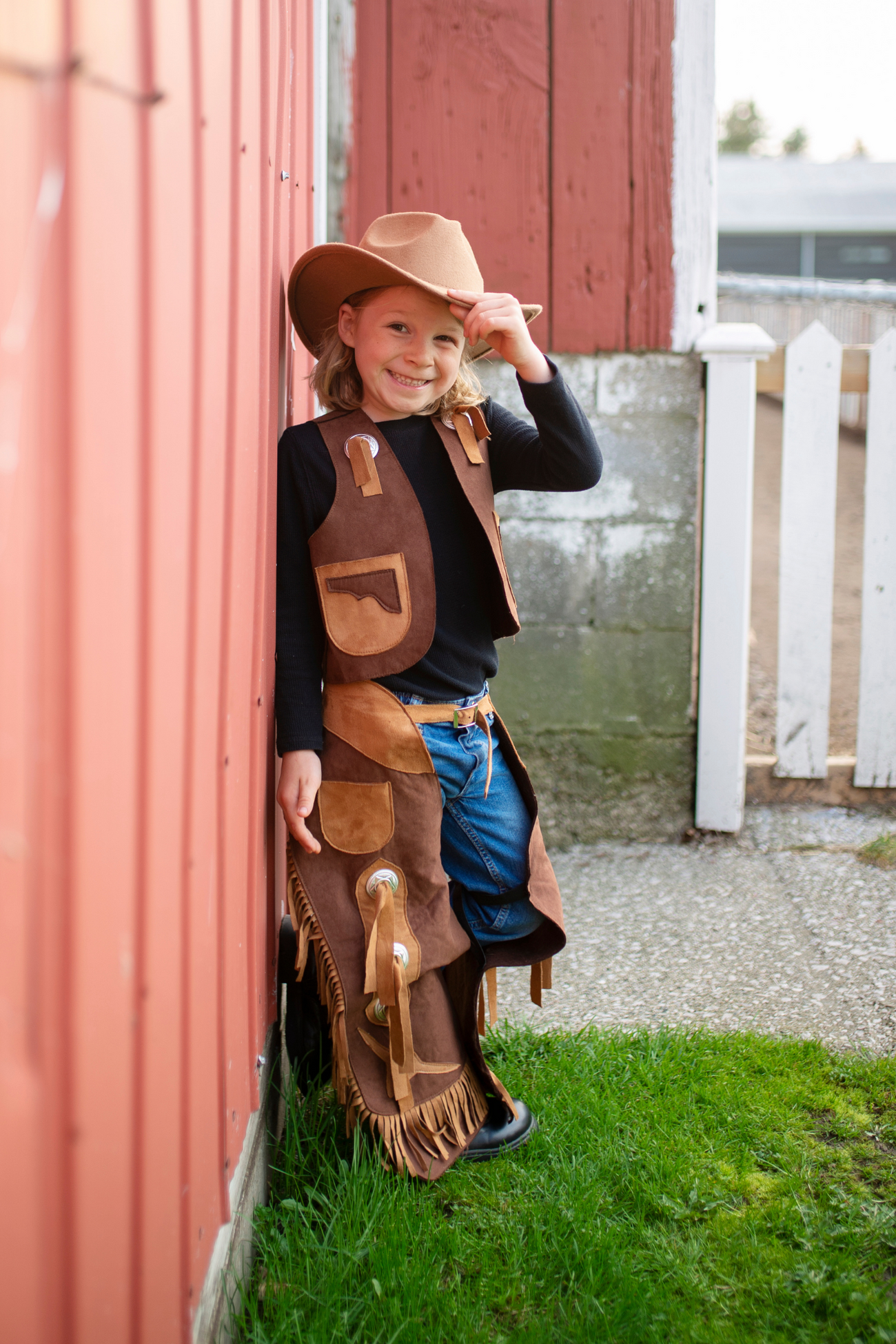 A child in a Great Pretenders Cowboy Vest and Chaps, size 7-8, smiles warmly by a red wooden wall, capturing the playful spirit of the Wild West.