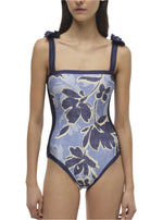 A person faces forward wearing Simkhai's Bow Strap One Piece swimsuit in Blue Hydrangea Floral with shoulder ties.