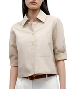 A woman wears the TWP Short Lived Top, a beige, short-sleeve cotton poplin button-up with a chest pocket and boxy fit, tucked into white pants and styled with a brown belt.