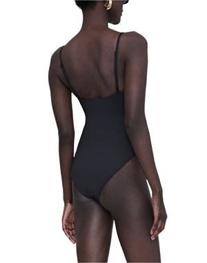 A person is seen from the back, standing with one arm relaxed at their side, wearing the Anine Bing Vesta One Piece—a black minimalist swimsuit with adjustable straps.