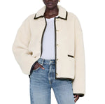 A person wearing the Anine Bing Camden Jacket—a cream wool blend sherpa workwear style with black trim—over a white t-shirt and blue jeans, stands with one hand in their pocket.