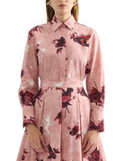 A woman wearing the Erdem Long Sleeve Abstract Rose Shirt, a pink cotton poplin collared dress with dark red floral print and pleated skirt, shown from the waist up.