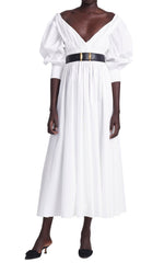 The person is wearing the Altuzarra Kathleen Optic White Dress, featuring a V-neck and puffed sleeves, cinched with a black belt, and paired with sleek black heels.