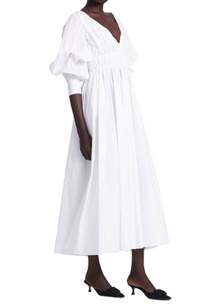 A person stands against a white background wearing the Kathleen Optic White Dress by Altuzarra, featuring a V-neck and puffed sleeves, complemented by black heeled shoes.