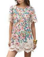 A woman wears the Figue April Dress, a short-sleeved cotton poplin floral-print dress with scalloped embroidery at the hem and sleeves in pink, green, and blue, accessorized with gold bracelets.