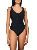 A woman models the Karla Colletto Basics V-Neck Tank with High Back One Piece, a black swimsuit featuring wide straps and center-front ruching for a flattering, shape-enhancing fit, set against a white background.