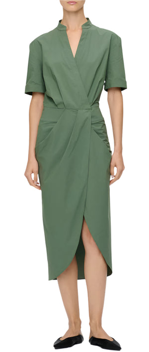 A person wearing the Callas Milano Bali Dress, a green, knee-length, short-sleeved wrap-style dress made from ethically sourced cotton, paired with black flat shoes.