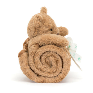 A brown plush Jellycat Bartholomew Bear Blankie sits atop a rolled-up soft tan blanket, tied with a white grosgrain ribbon and now crafted using recycled fibers for sustainability.