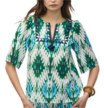 A woman wears the Figue Bodhi Top, a patterned green, blue, and white cotton voile blouse with elbow-length sleeves and delicate embroidery highlighting the geometric V-neck design.
