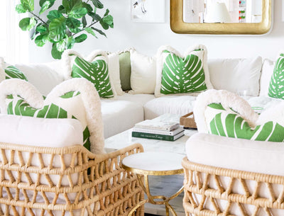 living room with white sofa, green and white pillows, fig leaf tree, gold mirror, and 2 woven cane chairs