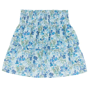 The Girls' Floral Skirt by Tartine et Chocolat, made from Liberty fabric, showcases a blue floral pattern on a white backdrop with a smocked elastic waist. Ideal for a breezy summer style.
