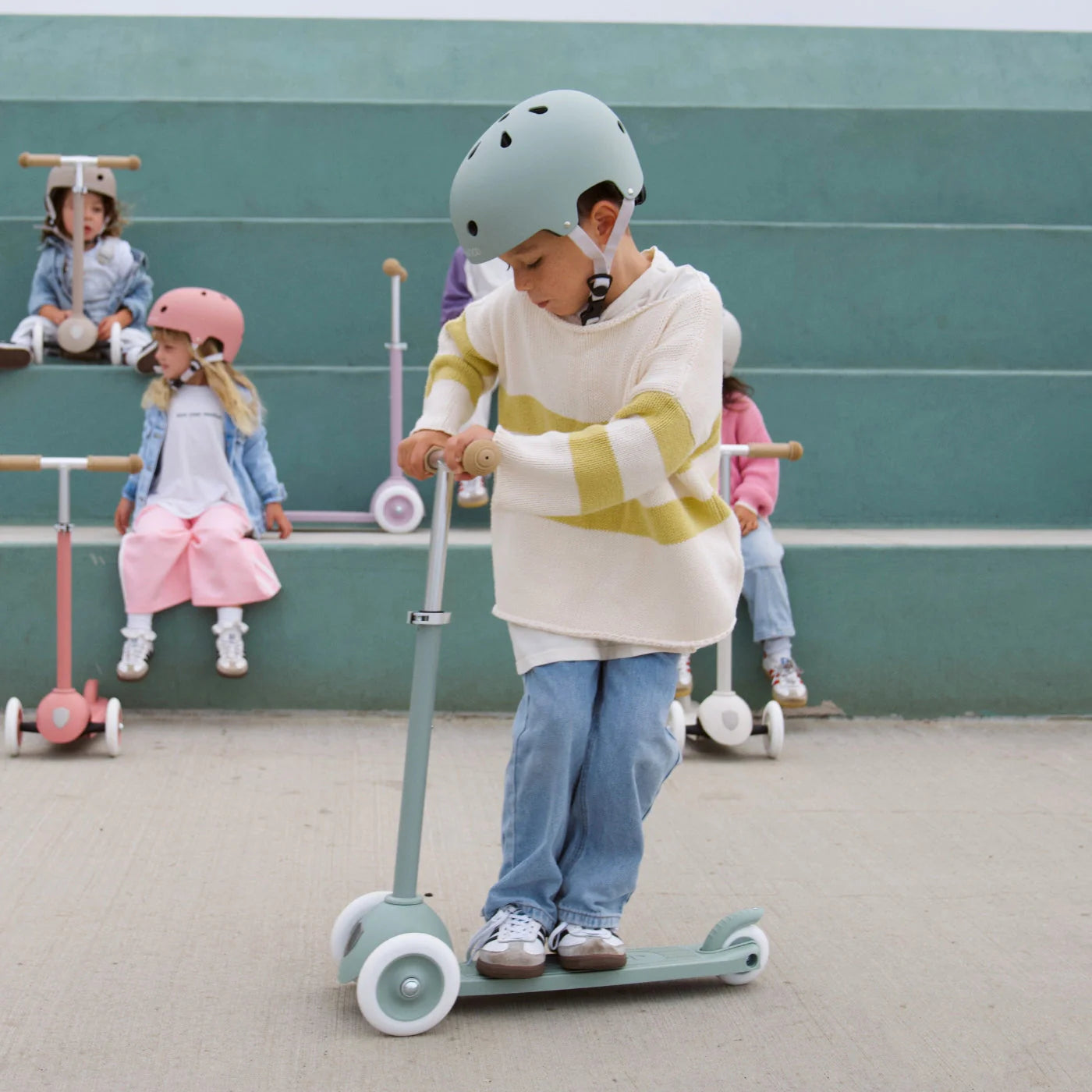 A child wearing a Banwood Eco Helmet rides a green scooter while other children in helmets watch from tiered green steps in the background.