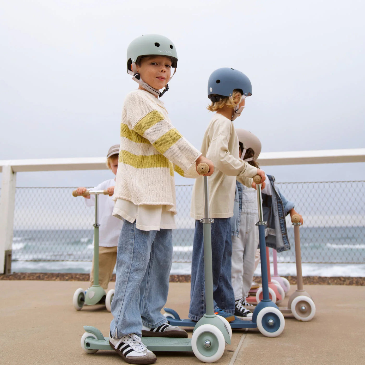Four children wearing helmets ride Banwood Eco Scooters from the Eco Collection along a paved path by the ocean, with a fence and waves in the background.