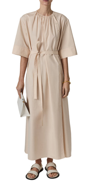 The individual is elegantly dressed in CO's Gathered Belted Dress, a long beige shirt dress with bell sleeves and a tied waist, paired with a small white bag. The ensemble is completed with white sandals and a stylish bracelet.