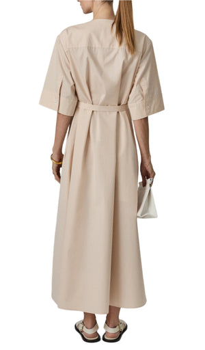A person with long hair wears CO's Gathered Belted Dress, an ankle-length beige shirt dress. They carry a white handbag and wear black and white shoes, viewed from behind.