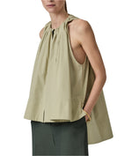 A person in a Gathered Tunic Shirt by CO, featuring a sleeveless light olive halter neck style paired with a dark green skirt, both crafted from soft cotton poplin, hands in pockets.