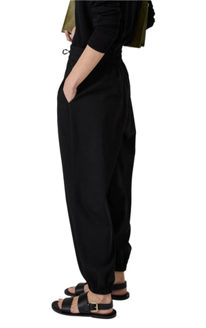 A person in CO's Jogger Pant, designed with black stretch wool-like fabric for a soft and comfortable fit, stands hands in pockets. The joggers showcase ankle cuffs and a tapered leg, paired with sandals.