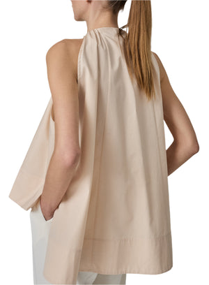 Someone with long hair tied back is wearing the Gathered Tunic Shirt, a sleeveless, loose-fitting trapeze top in beige cotton from CO, paired with white pants, viewed from the side and back.