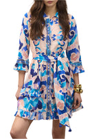 A woman wears the Figue Cami Dress, a knee-length dress with blue, pink, and white abstract prints, tie waist, ruffle details, tiered skirt; styled with gold bracelets.