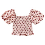 The Girls’ Trixie Top by Rylee & Cru features a smocked bodice, short puffed sleeves, pink cropped fit with red floral print, square neckline, and ruffled hem.