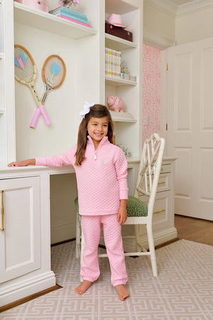 A young girl in Beaufort Bonnet Company Quilted Gates Sweeney Sweatpants stands in a white, neatly organized bedroom with a desk, chair, and tennis rackets on the wall.