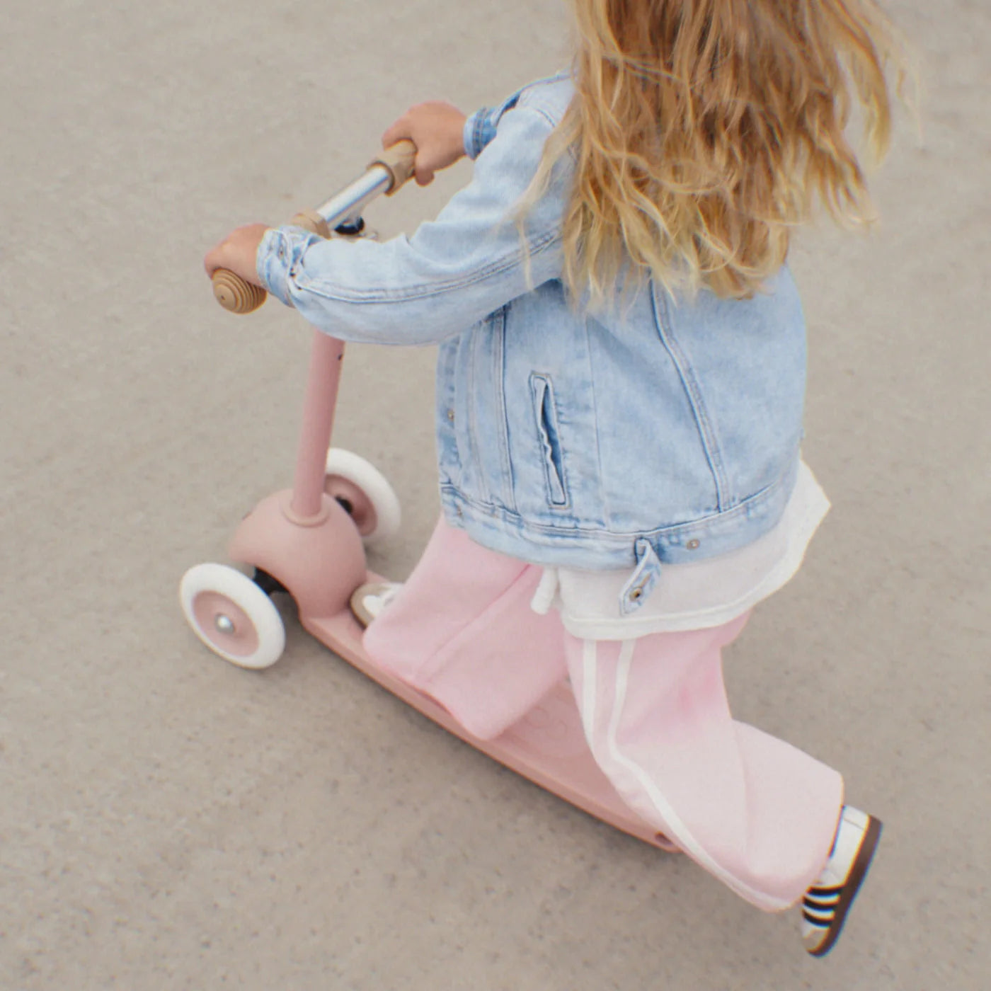 A child with long blonde hair, wearing a light blue denim jacket and pink pants, rides a pink Banwood Eco Scooter—crafted from recycled plastic—on a paved surface.