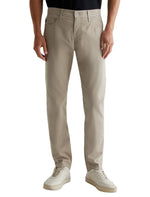 Dressed in AG Jeans Tellis Modern Slim Pant made from Italian sateen, paired with white sneakers, a person stands against a plain background.