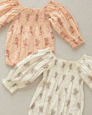 Two Noralee Baby Girls' Autumn Rompers with smocked empire bodice and bubbled long sleeves, one peach with red flowers, the other white with grey flowers, are laid flat on a textured beige surface.