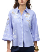 A woman wears the Figue Elaine Top, a light blue cotton poplin blouse with white floral embroidery, an embroidered front placket, three-quarter blouson sleeves, and lace cut-out details.