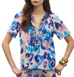 A woman models the Figue Elena Top, a short-sleeve cotton voile blouse in blue, pink, and white with custom embroidery at the neckline, paired with gold jewelry.