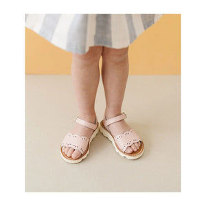 Ella, a cheerful spirit in a striped dress and her L'Amour Shoes Girls' Ella Scalloped Sandals, stands on a neutral floor against an orange background.
