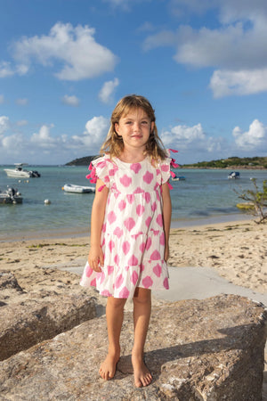 A young girl in the Mer Girls' Sophie Tassel Dress with a tiered skirt stands barefoot on a beachside rock, as boats float nearby under a partly cloudy sky.