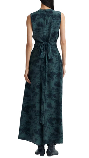 An individual wears the Altuzarra Penny Dress, featuring a sleeveless design with a nature pattern and tied waist sash. The long, dark green dress flows into a voluminous skirt elegantly covering black boots, viewed from the back.