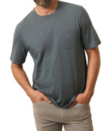 A man wears a short-sleeve, grayish-blue Faherty Sunwashed Pocket Tee made from Regenerative Organic Cotton and light gray pants, standing with his hands in his pockets.