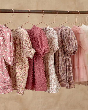 Six Noralee Girls' Louella Dresses in pink and mauve, featuring floral and patterned designs with puff sleeves and tiered skirts, hang on gold hangers from a rod against a beige background.