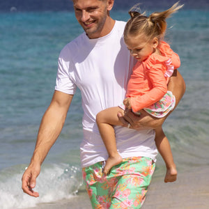 A man in a white shirt and floral swim trunks carries a young girl wearing the Snapper Rock Baby Girls' Coastal Shells Sustainable Ruffle Set along the shoreline, with ocean waves in the background.