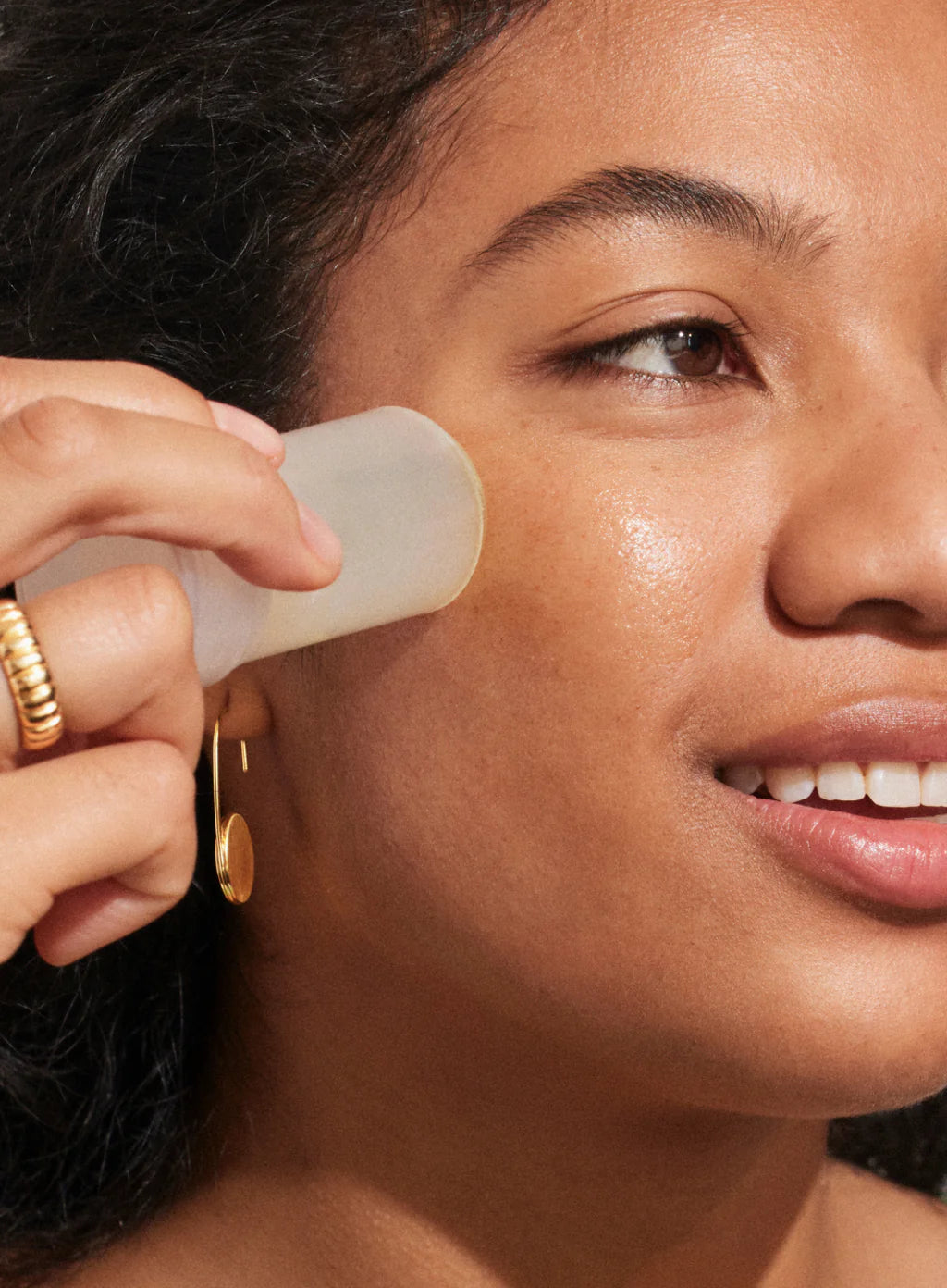 A person applies Supergoop Glow Stick SPF 50 to their cheek for a glowy finish, showcasing gold rings, a gold earring, and a radiant natural complexion.