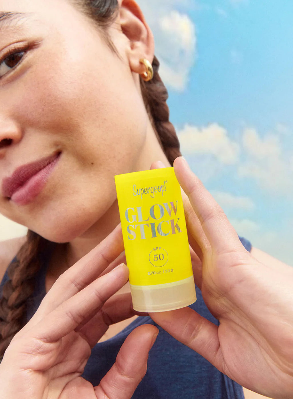 A person holds up a Supergoop Glow Stick SPF 50 sunscreen stick in front of their face against a blue sky, highlighting its portable design for an easy, radiant finish on the go.