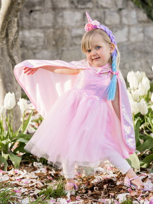 A young child in a pink unicorn costume, featuring a shimmering Sequins Cape by Great Pretenders with a velcro closure, delightfully stands among white tulips against the backdrop of a stone wall.